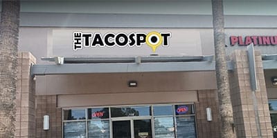 The Taco Spot restaurant exterior with "OPEN" signs in the windows, located between two palm trees.