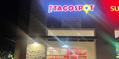 Street view of a restaurant at night displaying a lit neon sign that reads "The Taco Spot." The windows below feature additional signs, including an "Open Soon" notice.