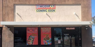 Exterior of a taco restaurant with "The Taco Spot Coming Soon" sign. The front windows display promotional images and social media information, with a "Welcome" sign on the door.