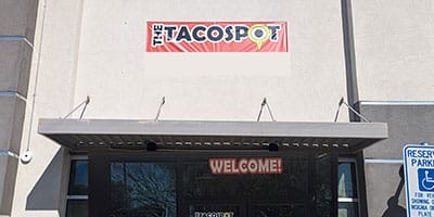 The image shows the exterior of The Taco Spot restaurant with a "WELCOME!" sign above the entrance. A reserved parking sign is visible on the right.