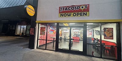 Street view of "The Taco Spot" restaurant. The storefront has a "Now Open" sign and large glass windows through which the interior dining area is visible.